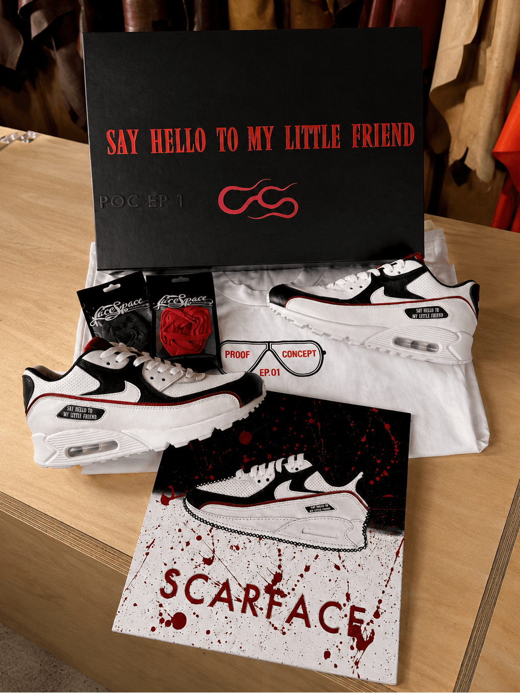A styled product display featuring a pair of black and white Nike sneakers with red accents, a black shoebox with red 'SAY HELLO TO MY LITTLE FRIEND' text, a white T-shirt, a small red rose, sunglasses, and a 'SCARFACE' card on a wooden table.