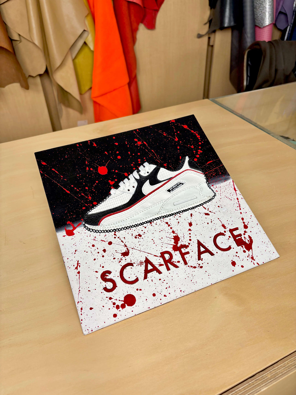 A square art print featuring a stylized white and black Nike sneaker on a black and white splattered background with the word 'SCARFACE' in red, displayed on a light wood table.
