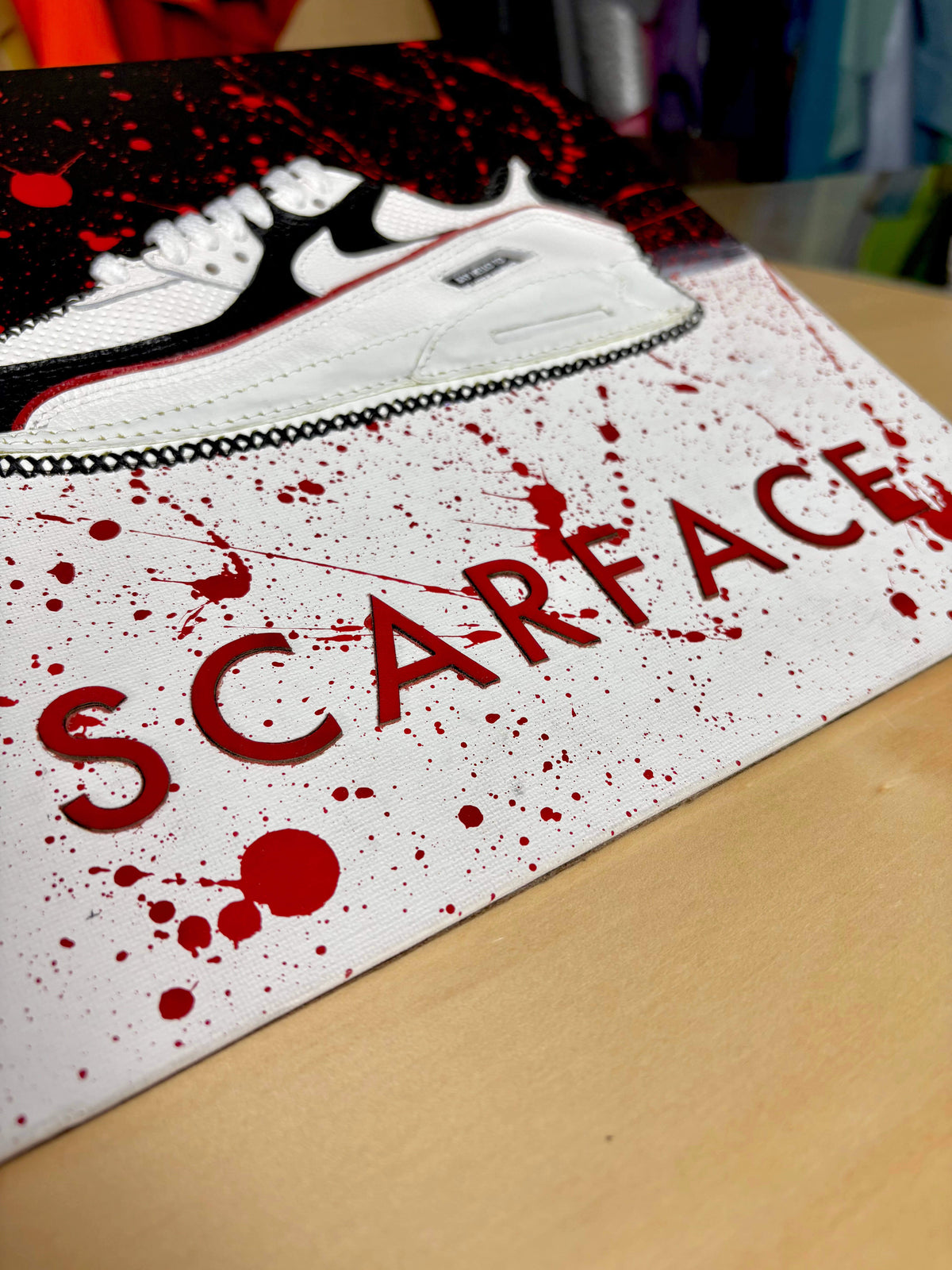 A close-up of a white sneaker with a black swoosh on a board printed with the word 'SCARFACE' in red letters and red blood-splatter graphics, resting on a light wood surface.