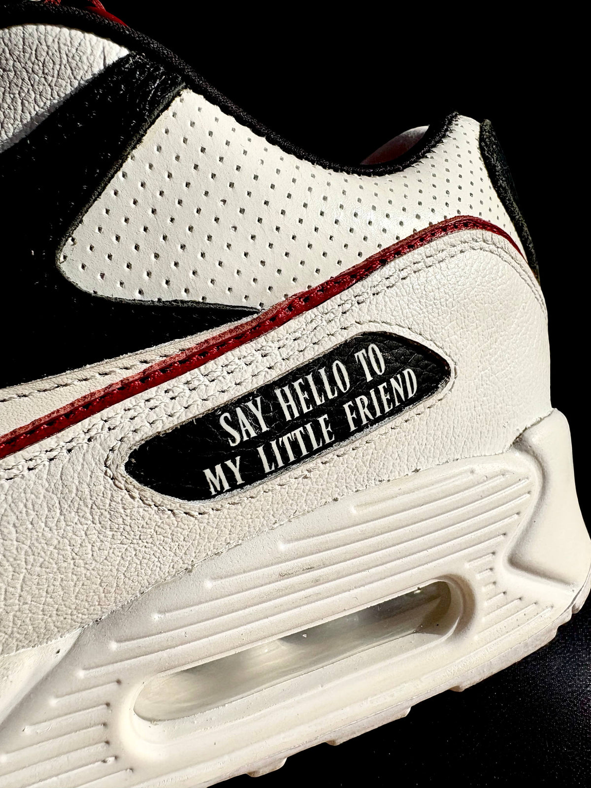 A close-up of a white leather sneaker with black overlays, red stitching, and a black patch that reads 'SAY HELLO TO MY LITTLE FRIEND'.