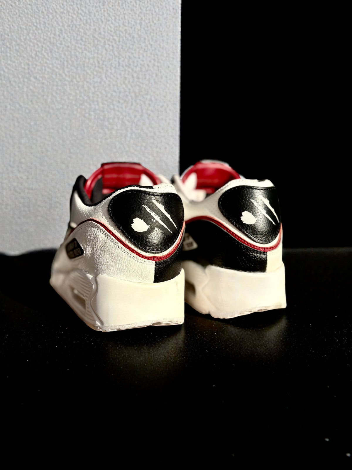 A pair of Nike Air Max 90 sneakers shown from the back, featuring white powder vinyl at the heel with black and white leather overlays, red accents, and white rubber soles.