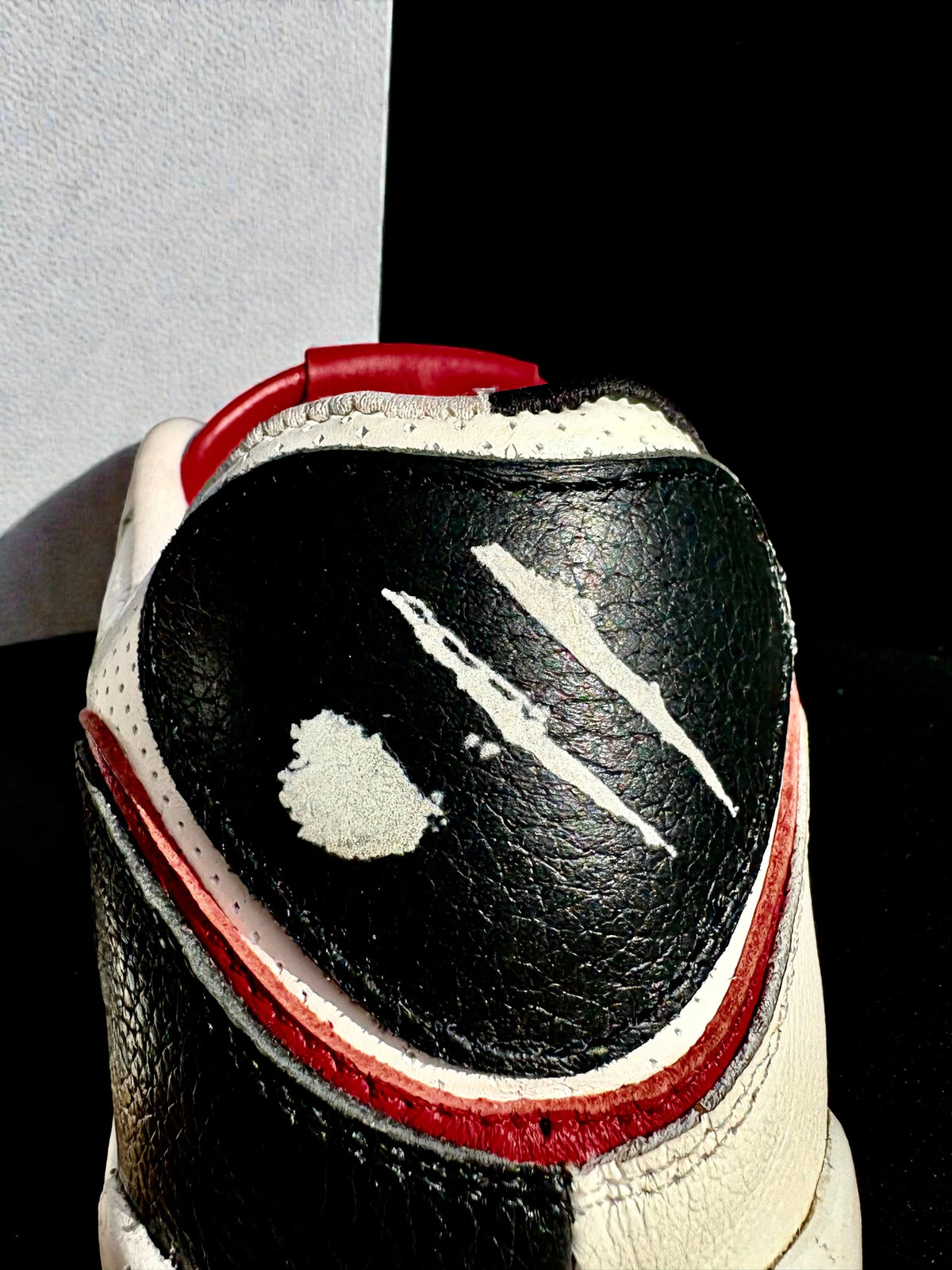 Close-up of the heel of a black, white, and red leather sneaker showing scuffed details, scratches, and a white vinyl splatter on the black leather panel.