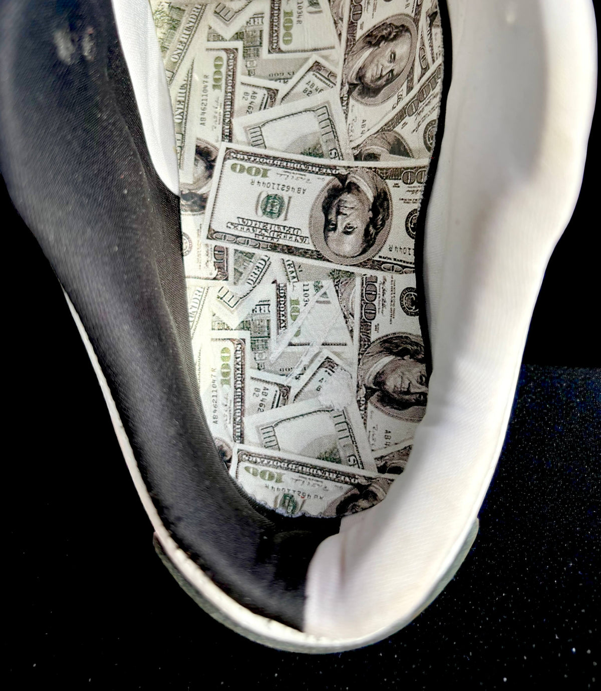 A close-up of a sneaker showing the black and white side panel and the white footbed printed with a collage of US $100 bill designs.